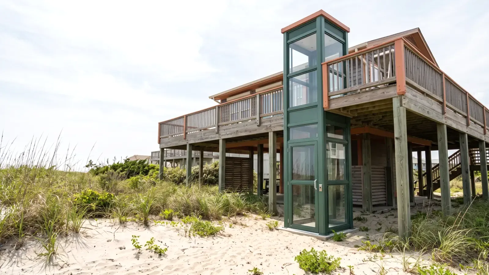 An outdoor home elevator tower with clear panels fixed beside a raised coastal house, reaching its upper deck